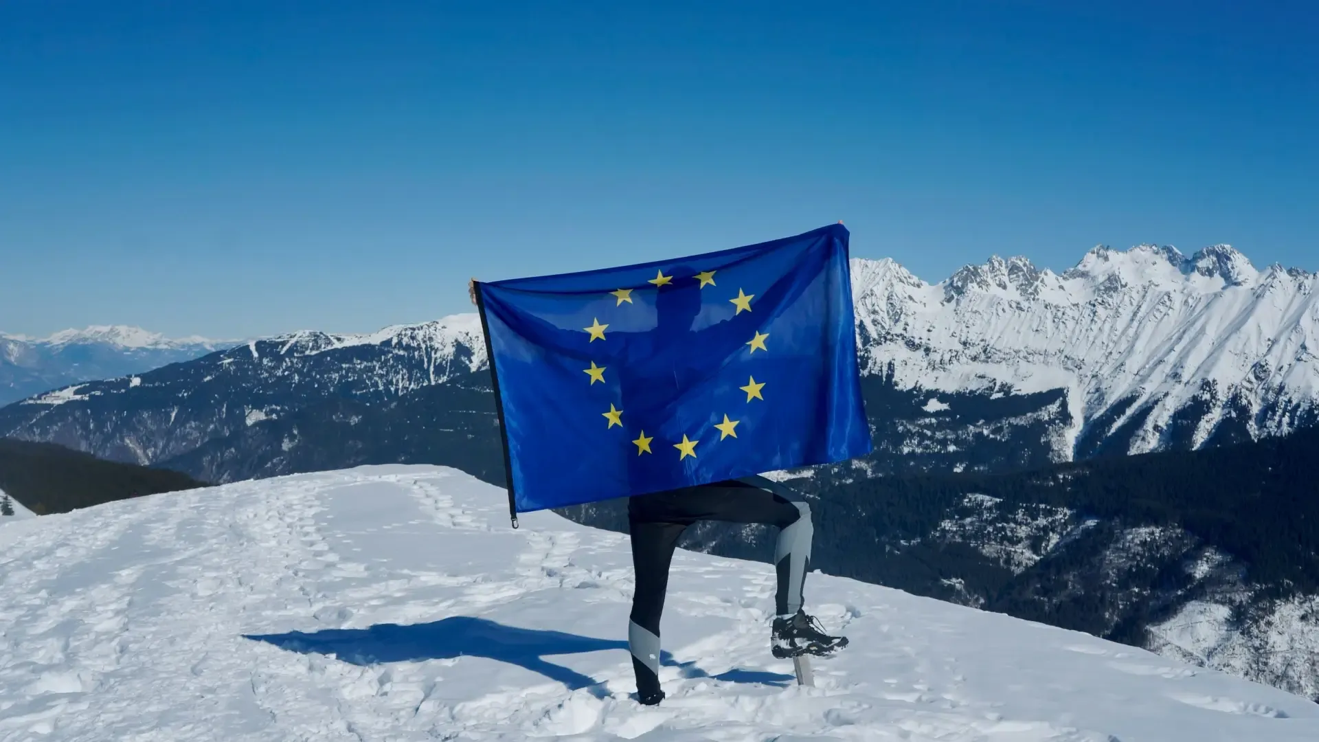A person holding the flag of the European Union atop of a snowy mountain peak